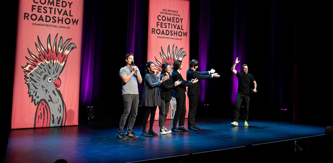 Image of five comedians on stage with two large banners behind them with the Melbourne International Comedy Festival Roadshow logo on them