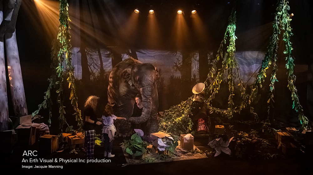 Image of a stage filled with jungle vines with a large puppet of an elephant at the rear. Three actors stand in front of the elephant talking.